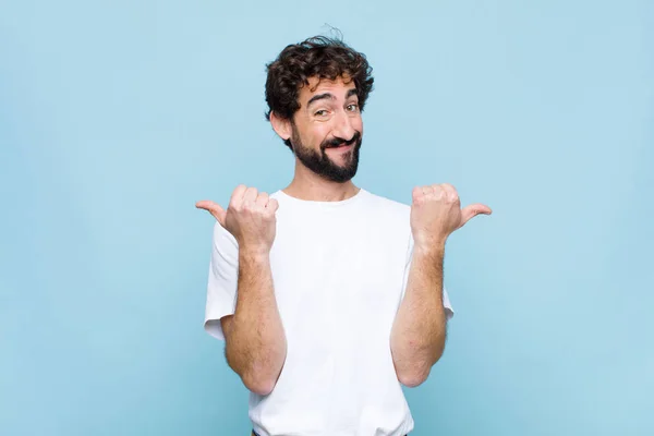 young crazy bearded man smiling joyfully and looking happy, feeling carefree and positive with both thumbs up against flat wall