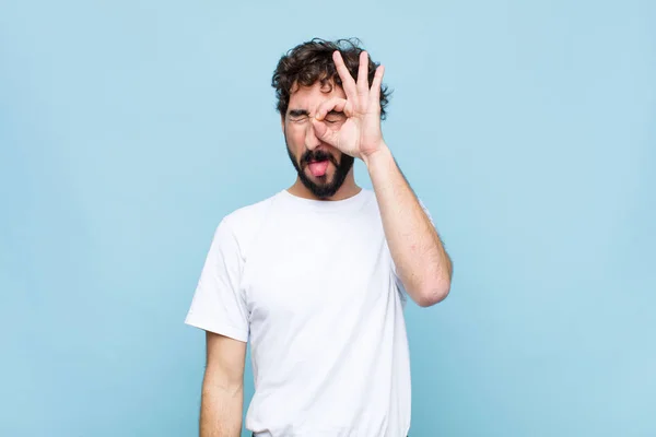 young crazy bearded man smiling happily with funny face, joking and looking through peephole, spying on secrets against flat wall