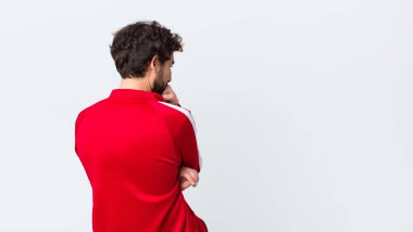 Genç sakallı bir adam ciddi, düşünceli ve endişeli görünüyor. Elleri çenesine bastırılmış bir şekilde fotokopi duvarına bakıyor.