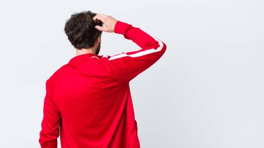 Genç sakallı adam düşünüyor ya da şüphe ediyor, kafasını kaşıyor, kafası karışık ve kafası karışık hissediyor, arka veya arka görünümü fotokopi duvarına karşı