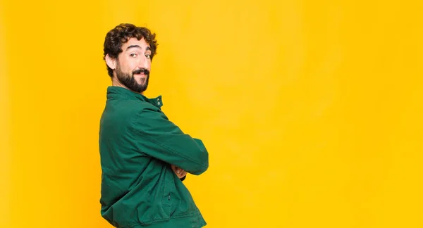 young crazy bearded man smiling gleefully, feeling happy, satisfied and relaxed, with crossed arms and looking to the side against flat wall