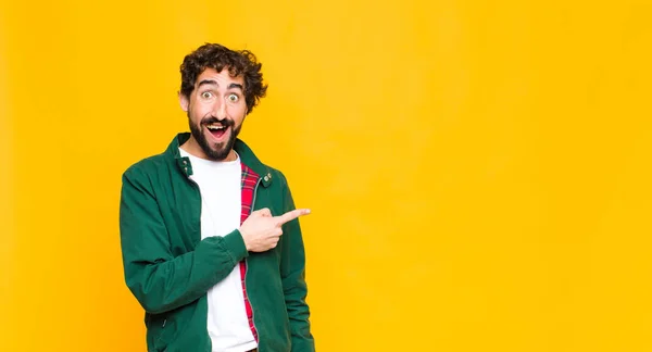 young crazy bearded man looking excited and surprised pointing to the side and upwards to copy space against flat wall
