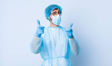 Olumlu, başarılı, mutlu bir tavırla gülümseyen genç adam kamerayı işaret ederek, elleriyle silah işareti yapıyor. Coronavirus konsepti