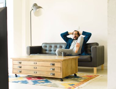 Oturma odasında kanepede oturup televizyon ya da bilgisayar izleyen genç havalı bir adam.