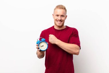 Genç sarışın adam mutlu, pozitif ve başarılı hissediyor, meydan okumayla karşılaştığında motive oluyor ya da alarm saati tutan beyaz duvara karşı iyi sonuçları kutluyor.