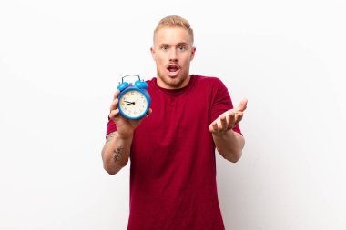 Genç sarışın adam şok olmuş, şaşırmış, endişeli ve paniklemiş hissediyor. Stresli ve dehşete kapılmış bir şekilde elinde çalar saatle beyaz duvara bakıyor.
