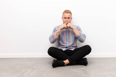 Genç sarışın adam ciddi ve hoşnutsuz görünüyor. Reddedilmiş olarak iki parmağıyla çimento zeminde oturarak sessizlik istiyor.