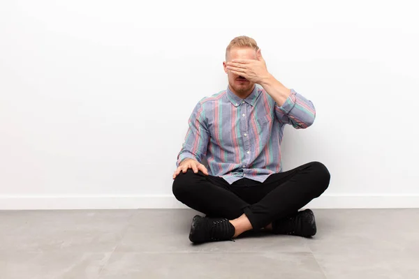 stock image young blonde man covering eyes with one hand feeling scared or anxious, wondering or blindly waiting for a surprise sitting on cement floor