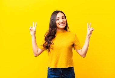 Genç Latin güzel kadın gülümsüyor ve mutlu görünüyor, arkadaş canlısı ve memnun, iki eli düz duvara karşı zafer ya da barış işareti yapıyor