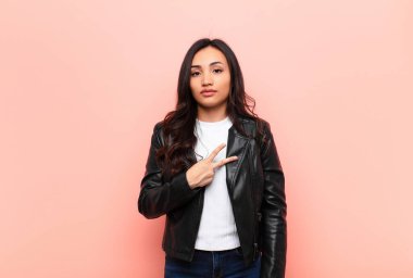 Genç Latin güzel kadın kendini mutlu, pozitif ve başarılı hissediyor. Göğsünde V şeklinde eller yapıyor. Düz duvara karşı zafer veya huzur gösteriyor.