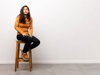 Genç ve güzel bir kadın şaşırmış ve şoka uğramış görünüyor, çenesi düşmüş, eli açık bir objeyi tutuyor ve bir odada oturuyor.