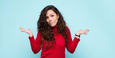 Genç ve güzel bir kadın şaşkın ve şaşkın hissediyor, şüpheci, farklı seçenekleri tartıyor ya da seçiyor mavi duvara karşı komik bir ifadeyle