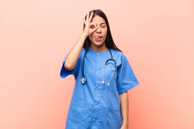 Genç Latin hemşire mutlu bir şekilde gülümsüyor, şaka yapıyor ve gözetleme deliğinden bakıyor, pembe duvara karşı sırları gözetliyor.