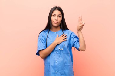 Genç Latin hemşire mutlu, kendinden emin ve güvenilir görünüyor. Gülümsüyor ve zafer işareti gösteriyor. Pembe duvara karşı olumlu bir tutum sergiliyor.