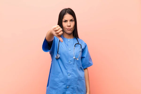 young latin nurse feeling cross, angry, annoyed, disappointed or displeased, showing thumbs down with a serious look against pink wall