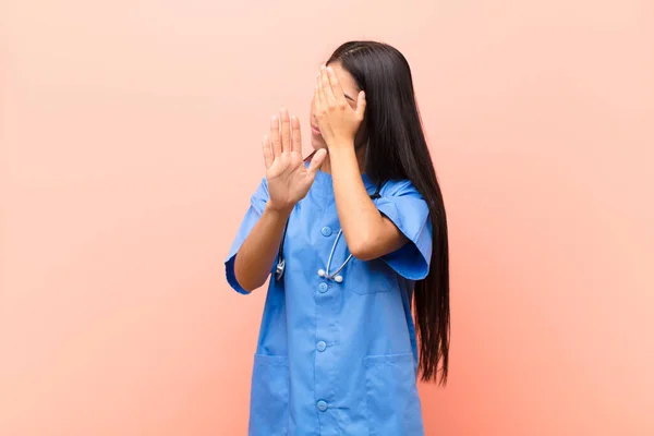 young latin nurse covering face with hand and putting other hand up front to stop camera, refusing photos or pictures against pink wall