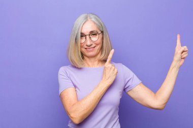 Yaşlı ya da orta yaşlı güzel bir kadın mutlu bir şekilde gülümsüyor ve iki elini de kopyalama boşluğunda nesne gösteriyor.