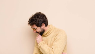 Genç, havalı bir adam mutlu, kendinden emin bir ifadeyle gülümsüyor, elleri çenesinde, merak ediyor ve düz duvara bakıyor.