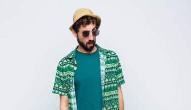 young tourist man feeling puzzled and confused, with a dumb, stunned expression looking at something unexpected against white wall