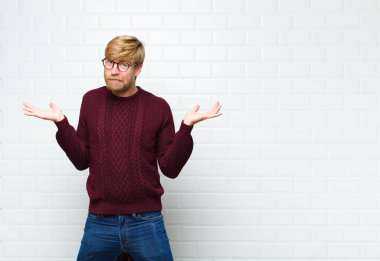 Genç sarışın adam şaşkın ve şaşkın hissediyor, şüpheci, farklı seçenekleri tartıyor ya da seçiyor. Komik ifadeli, klasik fayanslar duvarlı.