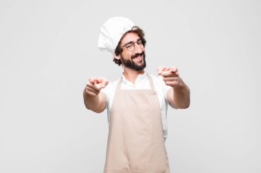 Genç çılgın şef mutlu ve kendinden emin hissediyor, kamerayı iki eliyle gösteriyor ve gülüyor, seni beyaz duvara karşı seçiyor.