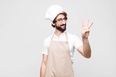 Genç çılgın şef gülümsüyor ve arkadaş canlısı görünüyor. Üç ya da üç numarayı gösteriyor. Elleri önde, beyaz duvara doğru sayıyor.