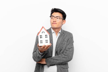 Genç Japon adam omuz silkiyor, kafası karışık ve emin değil, kolları çapraz ve şaşkın bir şekilde ev modeline bakıyor.