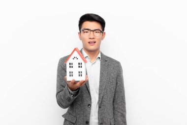 young japanese man feeling puzzled and confused, with a dumb, stunned expression looking at something unexpected with a house model