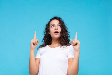 Genç ve güzel bir kadın şaşırmış, şaşırmış ve ağzı açık görünüyor, iki eliyle yukarıya doğru uzayı mavi arka plana kopyalamak için işaret ediyor.