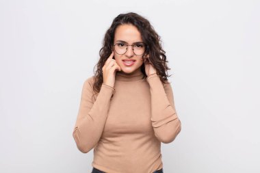 Genç, güzel bir kadın kızgın, stresli ve sinirli görünüyor, her iki kulağı da sağır edici bir gürültü, ses veya yüksek sesle kaplayan beyaz duvara karşı müzik