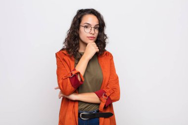 Genç, güzel bir kadın ciddi görünüyor, kafası karışık, kararsız ve düşünceli, beyaz duvara karşı seçenekler veya seçimler arasında şüpheci.