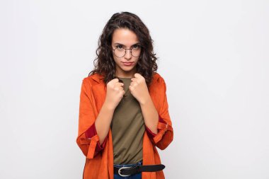 Genç, güzel bir kadın kendinden emin, sinirli, güçlü ve agresif görünüyor. Yumrukları beyaz duvara karşı boks pozisyonunda dövüşmeye hazır.