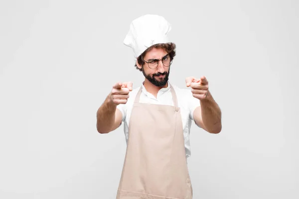 young crazy chef pointing forward at camera with both fingers and angry expression, telling you to do your duty against white wall