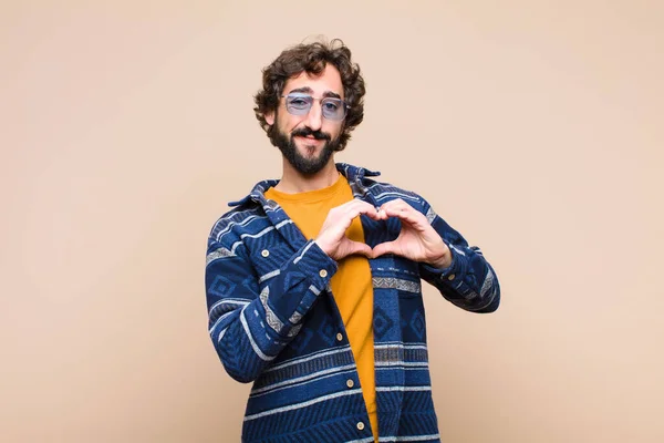 young crazy cool man smiling and feeling happy, cute, romantic and in love, making heart shape with both hands against flat wall