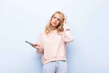 Genç sarışın kadın şaşkın ve kafası karışmış hissediyor. Kafasını kaşıyor ve akıllı telefon kullanıyor.