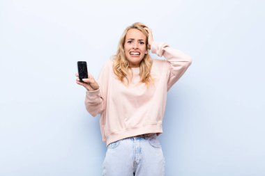 Genç sarışın kadın kendini stresli, endişeli, endişeli ya da korkmuş hissediyor, elleri başının üstünde, akıllı telefon kullanarak hata yaptığında panikliyor.