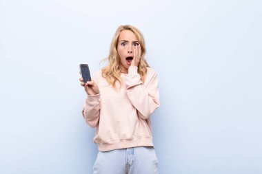 Genç sarışın kadın şok olmuş ve korkmuş hissediyor, korkmuş görünüyor ağzı açık ve elleri yanaklarında akıllı telefon kullanıyor.