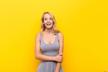 Genç sarışın kadın utangaç ve neşeli bir şekilde gülüyor, arkadaş canlısı ve olumlu ama turuncu duvara karşı güvensiz bir tutum sergiliyor.