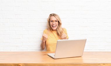 Genç sarışın kadın muzaffer bir şekilde bağırıyor, gülüyor, mutlu ve heyecanlı bir şekilde dizüstü bilgisayar kullanarak başarıyı kutluyor.