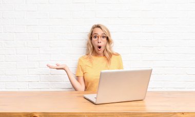 Genç sarışın kadın şaşırmış ve şok olmuş görünüyor, çenesi düşmüş, eli açık bir objeyi dizüstü bilgisayar kullanarak tutuyor.