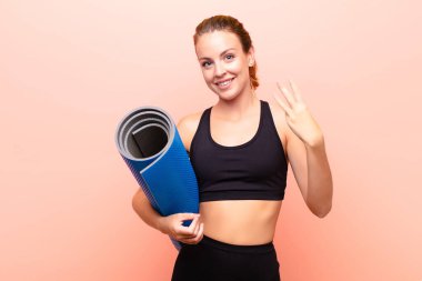 Kızıl saçlı güzel bir kadın gülümsüyor ve arkadaş canlısı görünüyor, üç ya da üçüncüyü gösteriyor, elinde yoga minderiyle geriye doğru sayıyor..