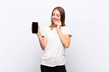 kızıl saçlı güzel bir kadın mutlu, kendinden emin bir ifadeyle gülümsüyor elleri çenesinde, merak ediyor ve elinde akıllı bir telefonla yan tarafa bakıyor.