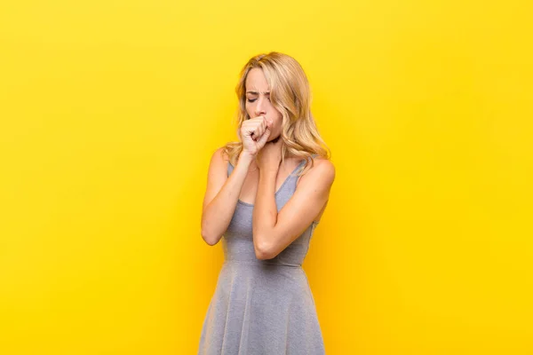 young blonde woman feeling ill with a sore throat and flu symptoms, coughing with mouth covered against orange wall