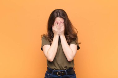 Genç, güzel bir kadın üzgün, hayal kırıklığına uğramış, gergin ve depresif, iki eliyle yüzünü kapatmış, turuncu duvara karşı ağlıyor.