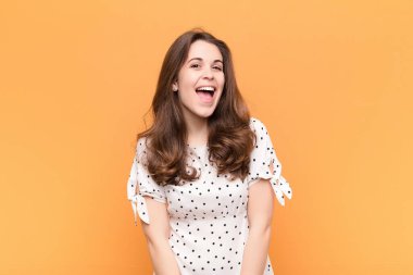 Büyük, dostane, kaygısız bir gülümsemesi olan genç, güzel bir kadın. Olumlu, rahat ve mutlu görünüyor. Turuncu duvarda takılıyor.