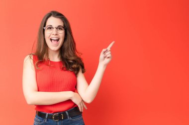 Genç ve güzel bir kadın mutlu bir şekilde gülümsüyor ve yan bakıyor, merak ediyor, düşünüyor ya da kırmızı duvara karşı bir fikri var.