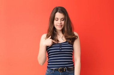 Genç ve güzel bir kadın neşeyle ve gelişigüzel gülümsüyor, aşağıya bakıyor ve göğsünü kırmızı duvara gösteriyor.