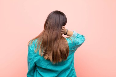 Genç güzel bir kadın düşünüyor ya da şüphe ediyor, kafasını kaşıyor, şaşkın ve şaşkın hissediyor, pembe duvarın arkasında ya da arkasında.