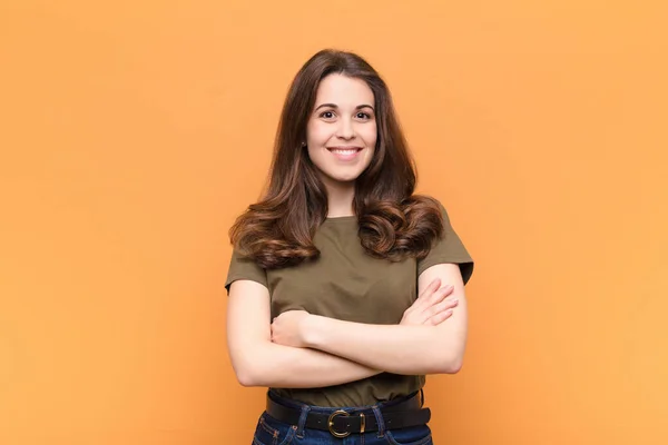 young pretty woman smiling to camera with crossed arms and a happy, confident, satisfied expression, lateral view against orange wall