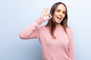 Genç, güzel bir kadın kendini başarılı ve tatmin olmuş hissediyor, ağzı açık gülümsüyor, elini mavi duvara yaslayarak işaret diliyle işaret veriyor.